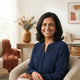 A professional portrait headshot of a smiling woman in a home environment with warm, neutral decor.