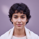 Close-up portrait of a smiling woman with short, dark, curly hair against a soft lilac background.